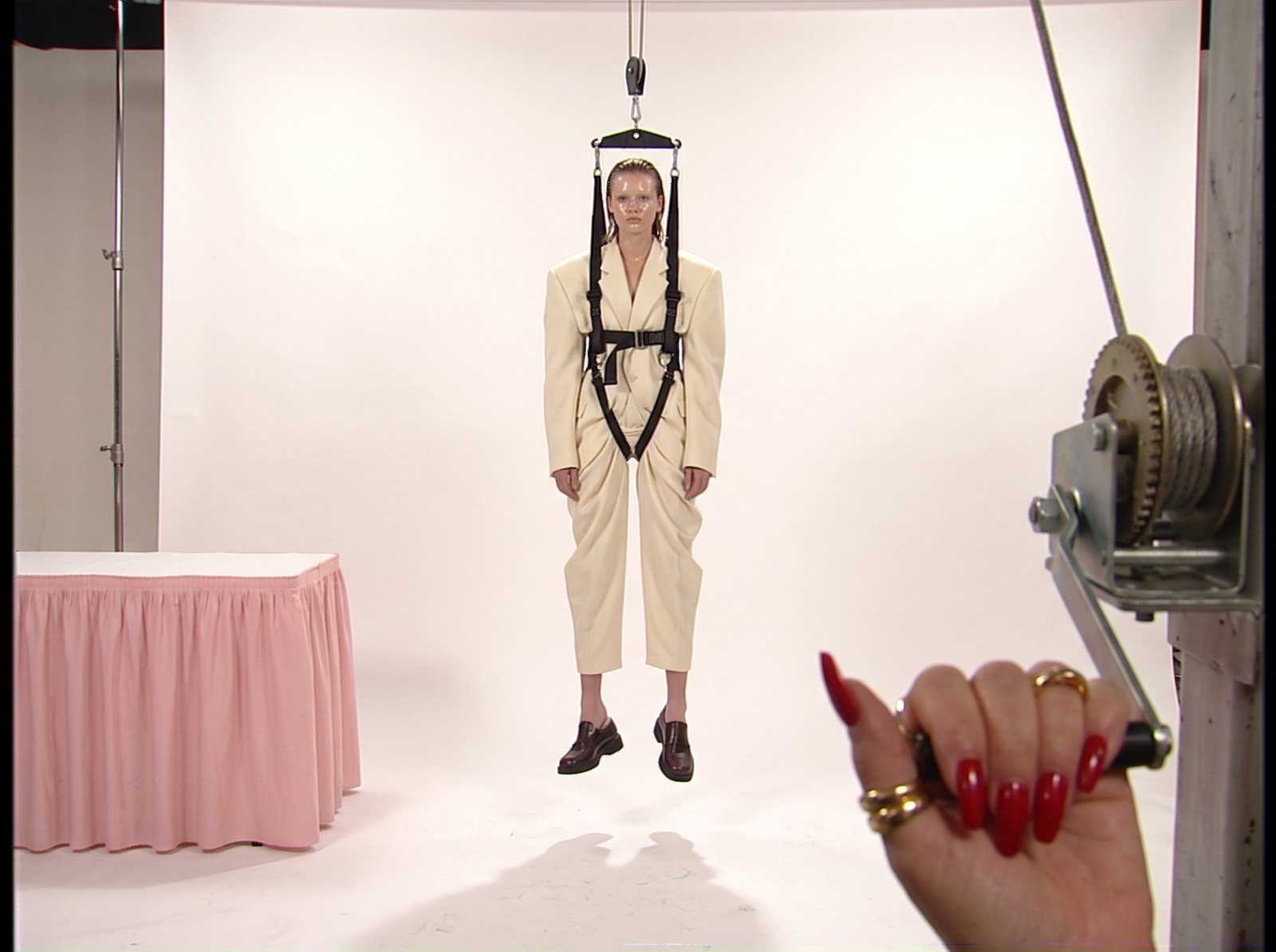 Style frame, recast: a model with slicked hair in a cream suit hangs in the harness while a hand with red nails and gold rings turns the winch
