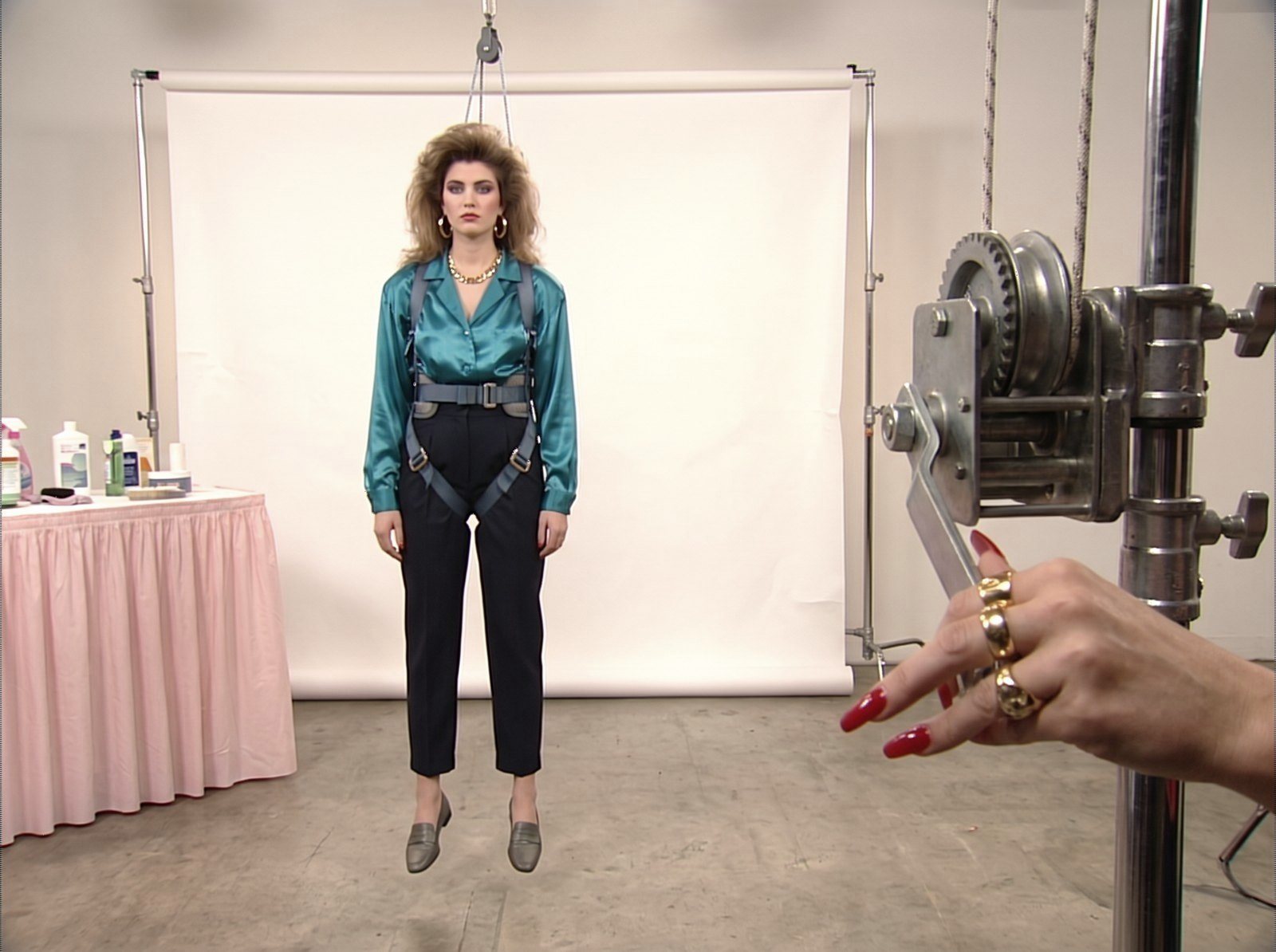 Style frame, all 90s: a model with big curled hair in a teal satin blouse stands in a harness by a winch on a white set