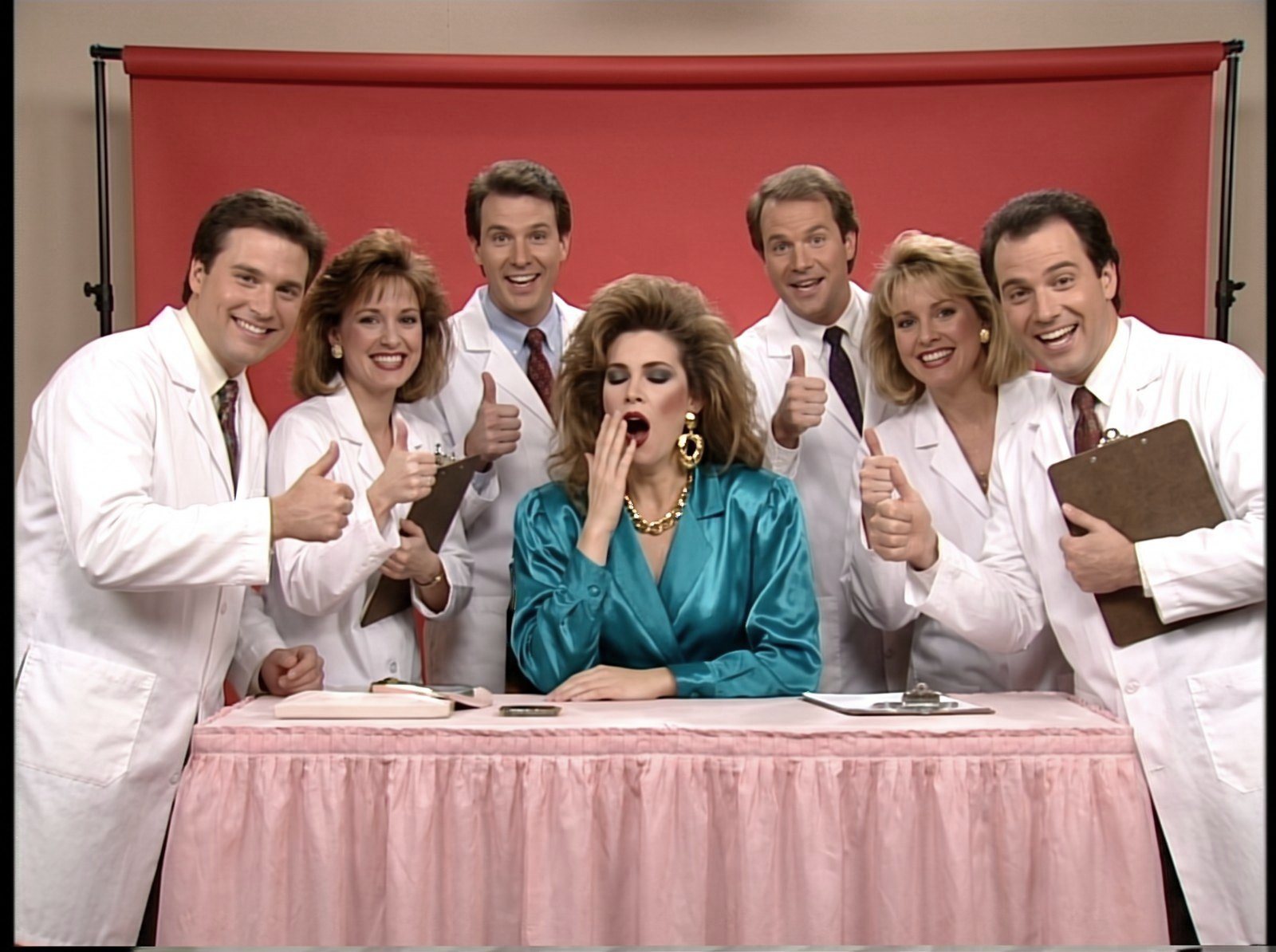 Style frame, all 90s: lab coats with feathered hair give a thumbs up around a yawning model in a teal satin blouse at a pink skirted table