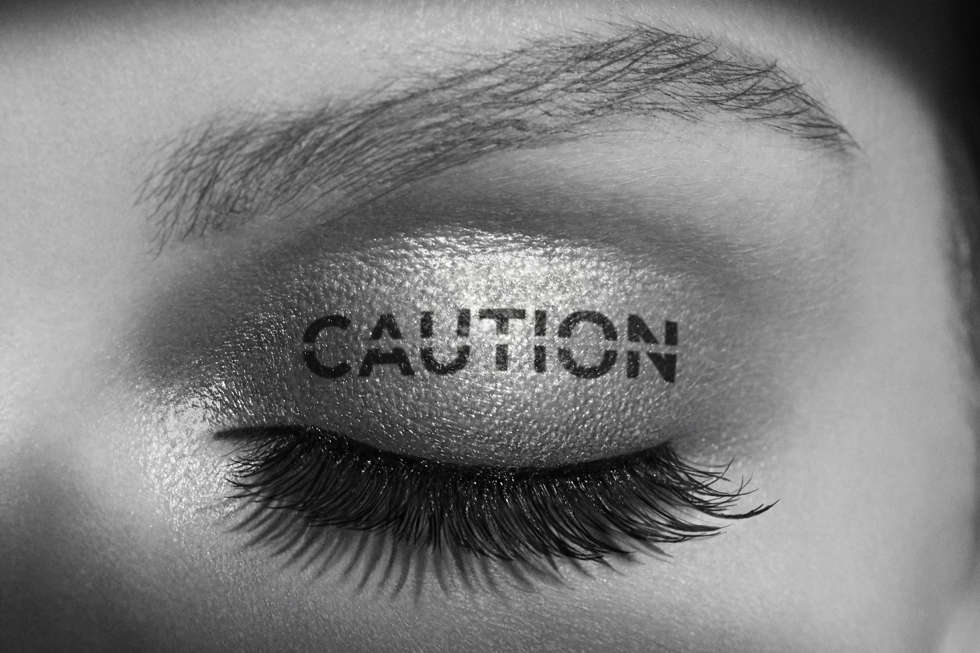 Close-up of a closed eye with long lashes, CAUTION stenciled across the shimmering lid