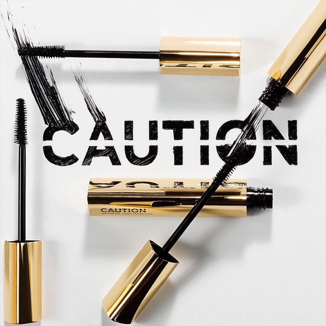Instagram post: CAUTION written in mascara strokes with gold components on white