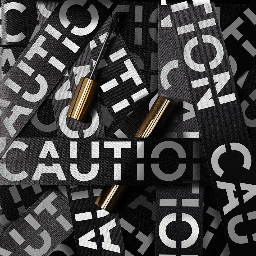 Instagram post: gold CAUTION mascaras among cut strips of CAUTION type