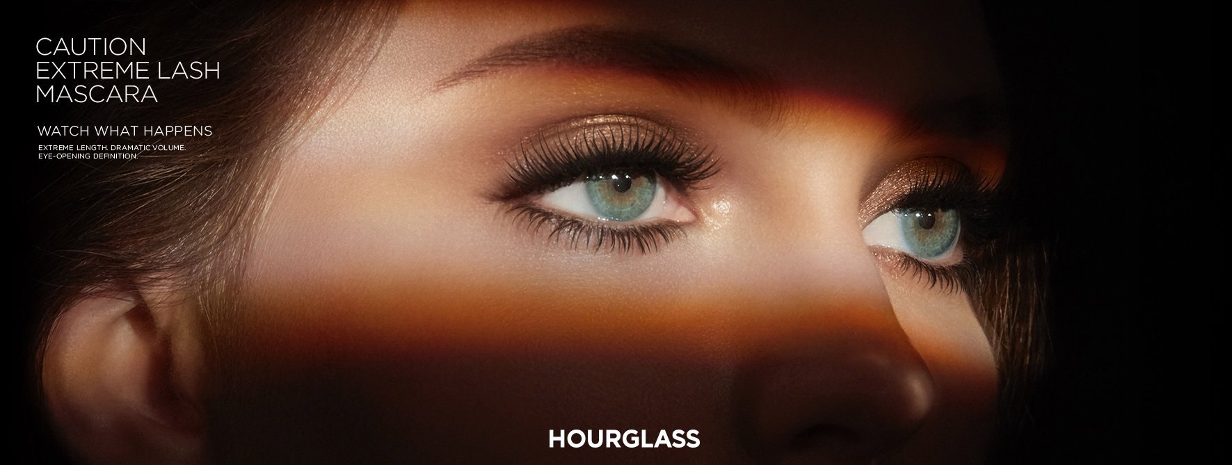 Wide banner: a woman’s green eyes in a band of warm light, CAUTION EXTREME LASH MASCARA and HOURGLASS in white
