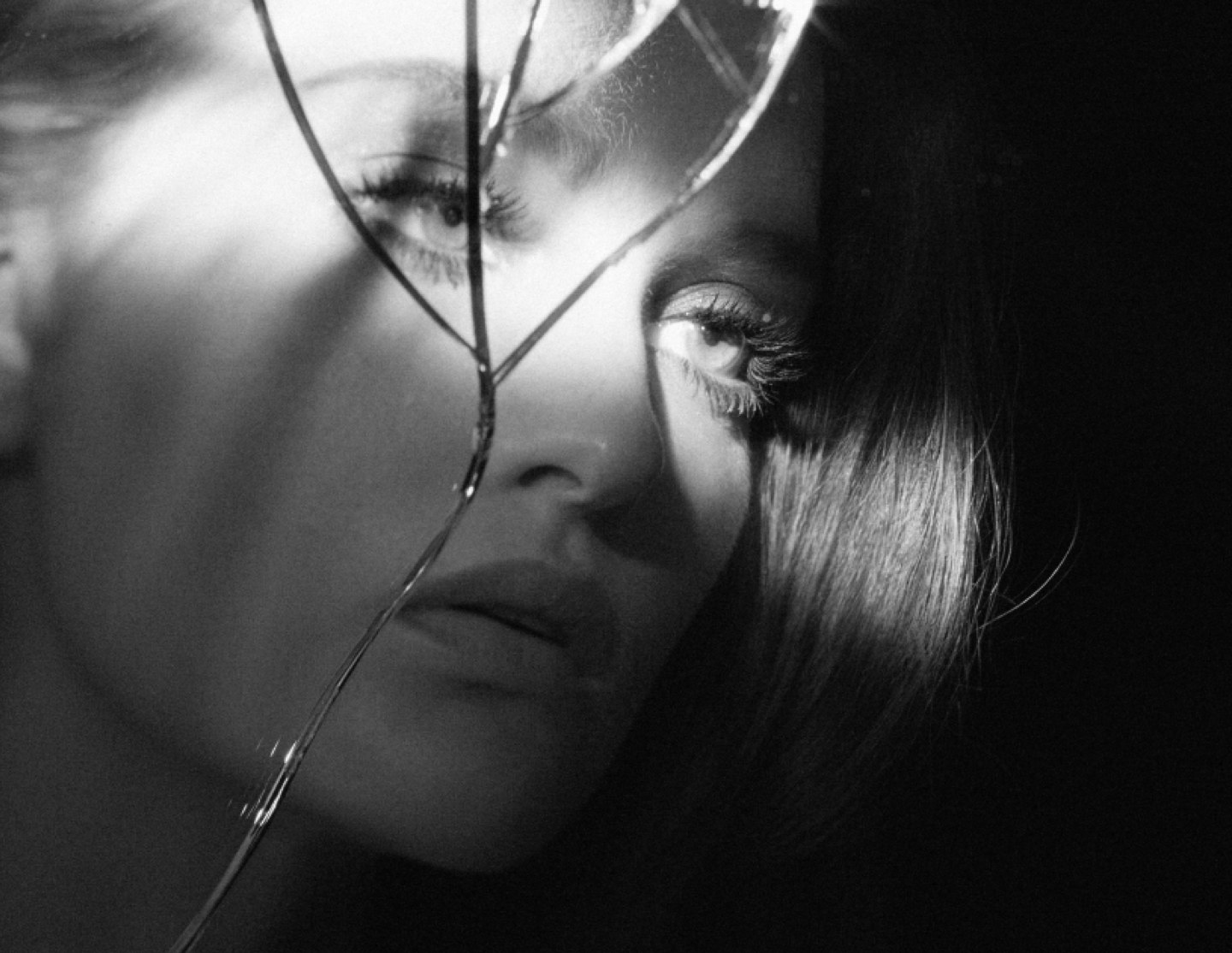 A woman’s face in black and white behind a cracked mirror, light falling across her eye and lashes