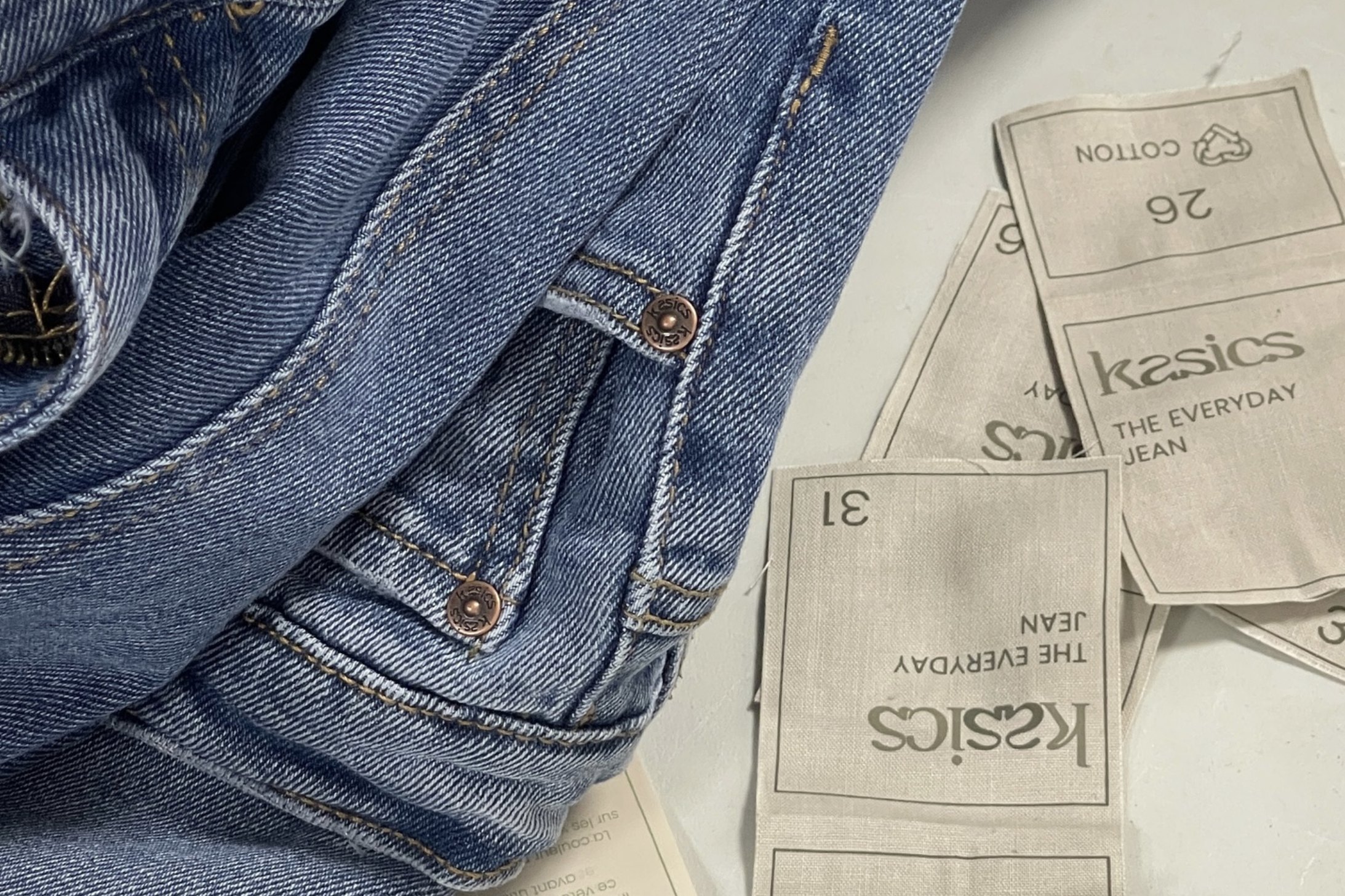 Woven kasics labels for The Everyday Jean beside folded denim with kasics stamped rivets