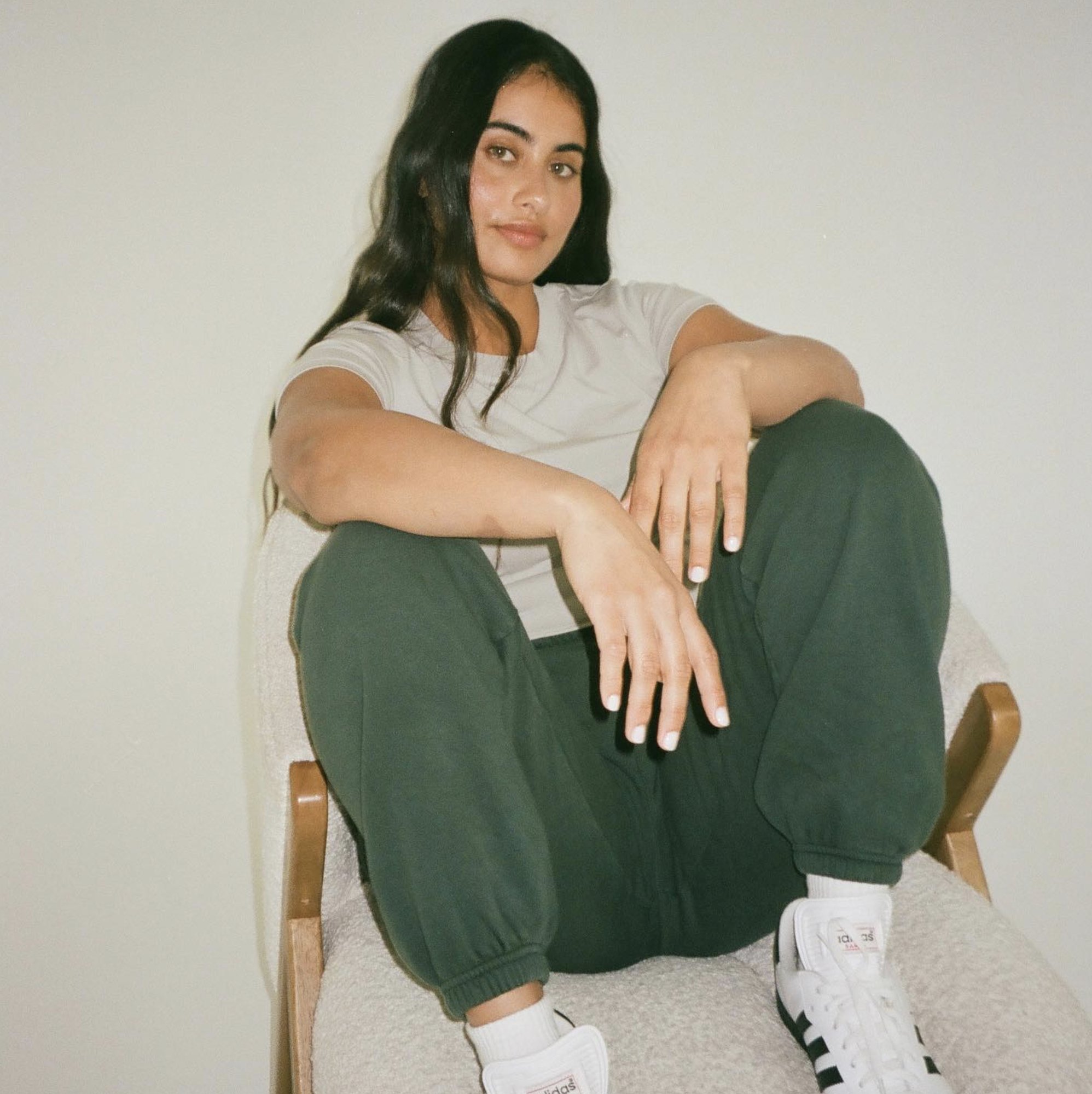 A woman in a pale grey tee and dark green sweatpants, sitting on a boucle chair against a white wall