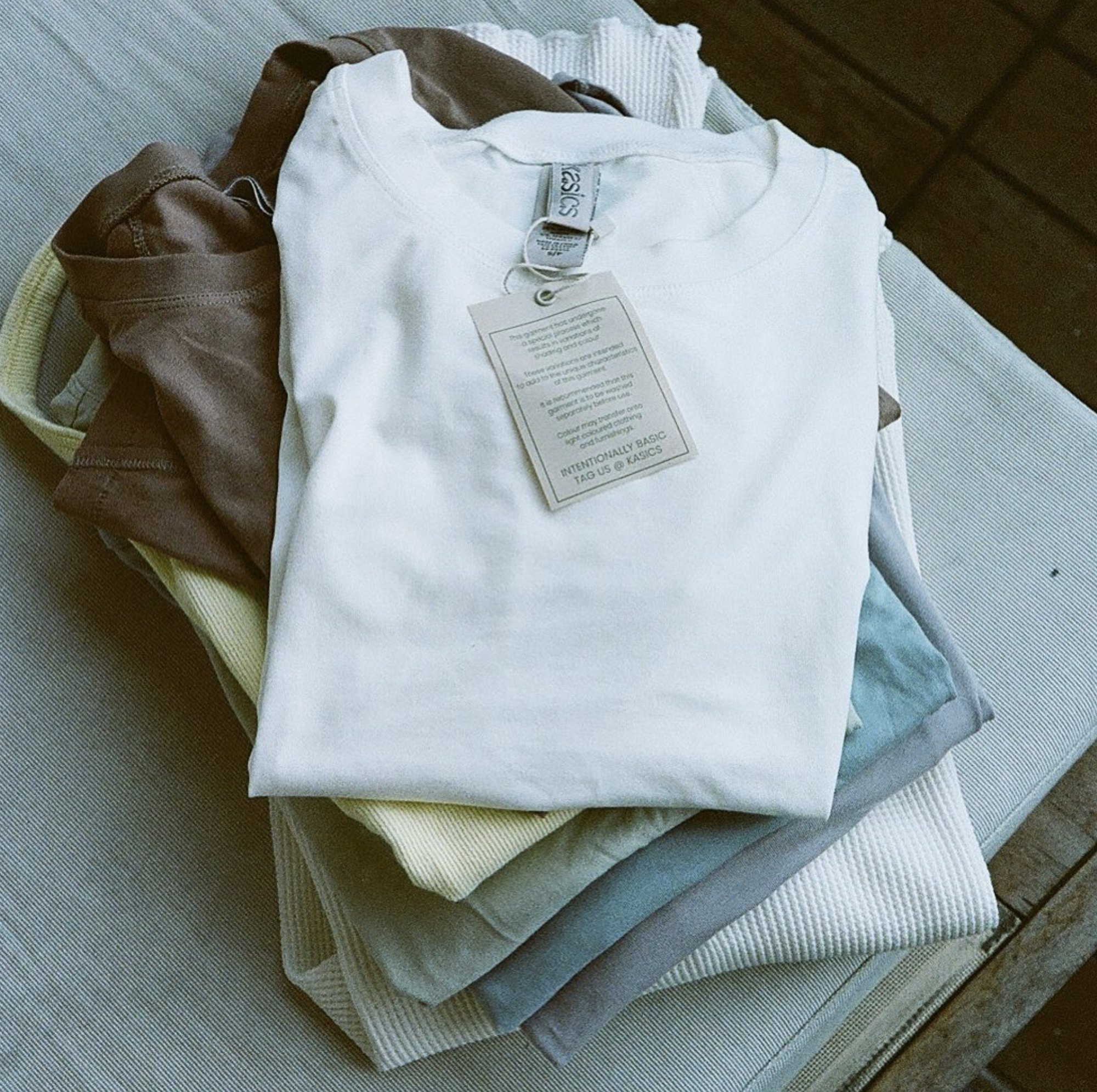 A stack of folded Kasics tees, the top one with a kasics neck label and a hang tag that reads Intentionally basic, tag us at Kasics