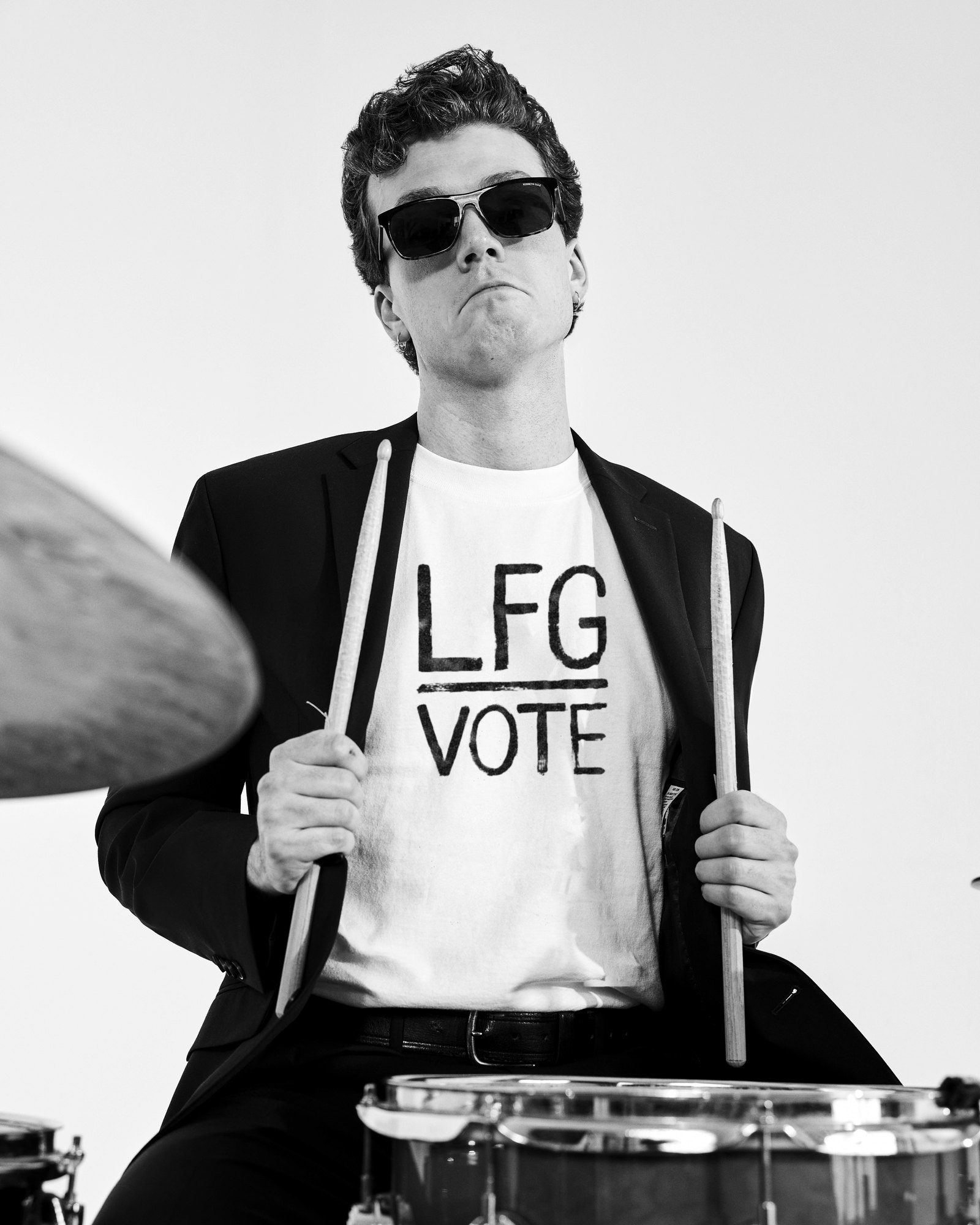 A drummer in a black suit and sunglasses holding his sticks, wearing the white LFG Vote tee, in black and white