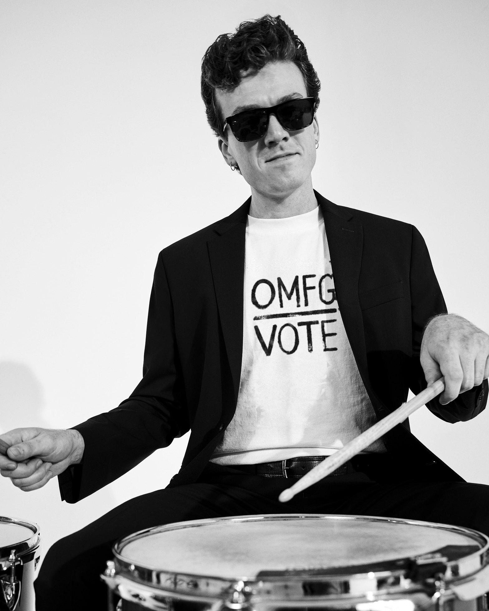 A drummer in a black suit and sunglasses playing the snare, wearing the white OMFG Vote tee, in black and white