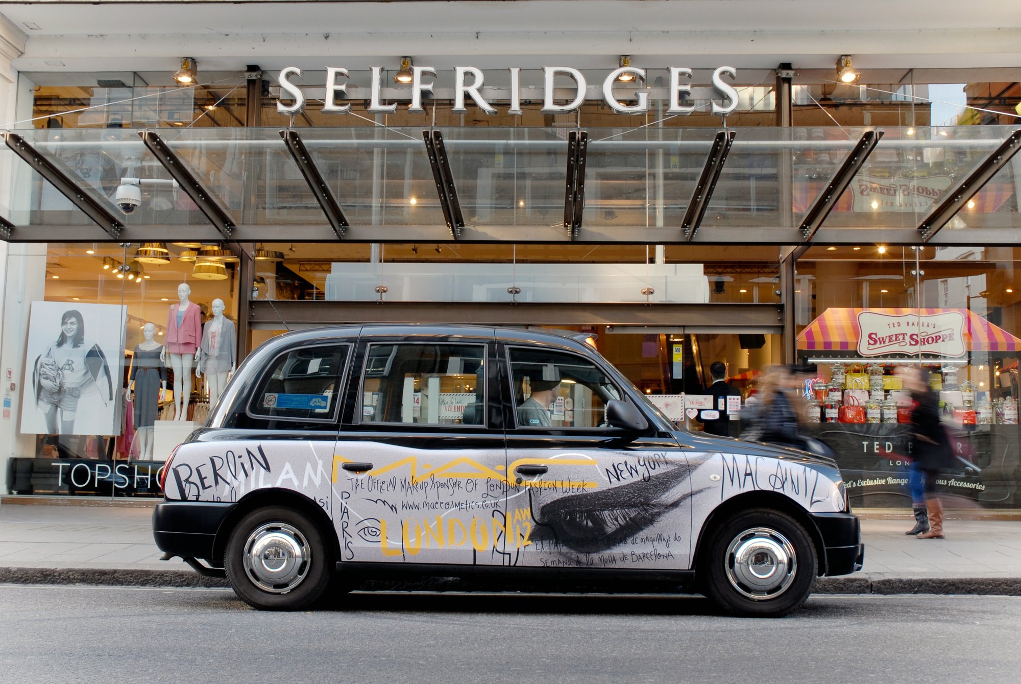 A London taxi wrapped in hand-lettered city names outside Selfridges