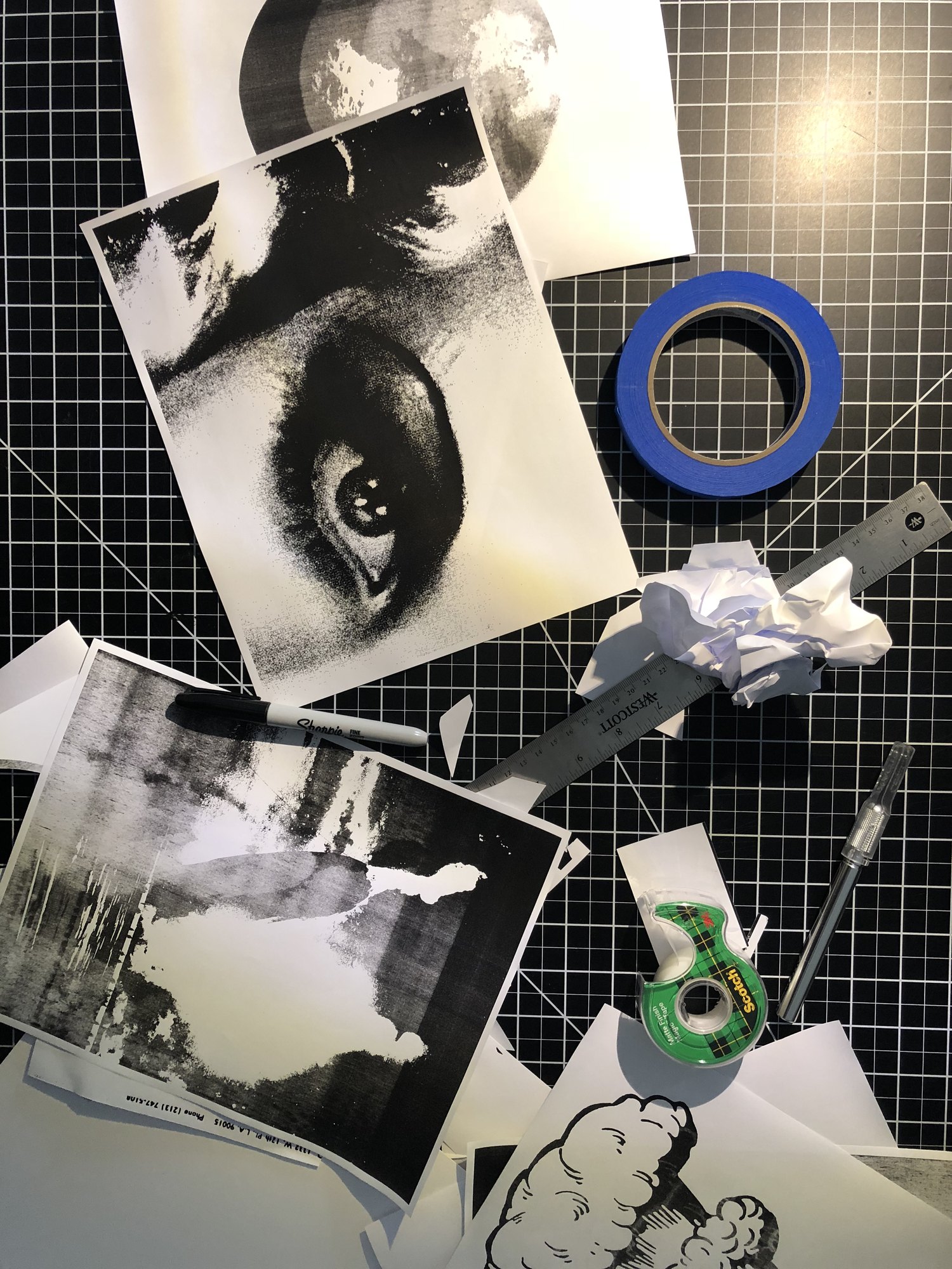 Xerox copies of a face and a cloud on a cutting mat with tape, a craft knife, a steel ruler and a marker