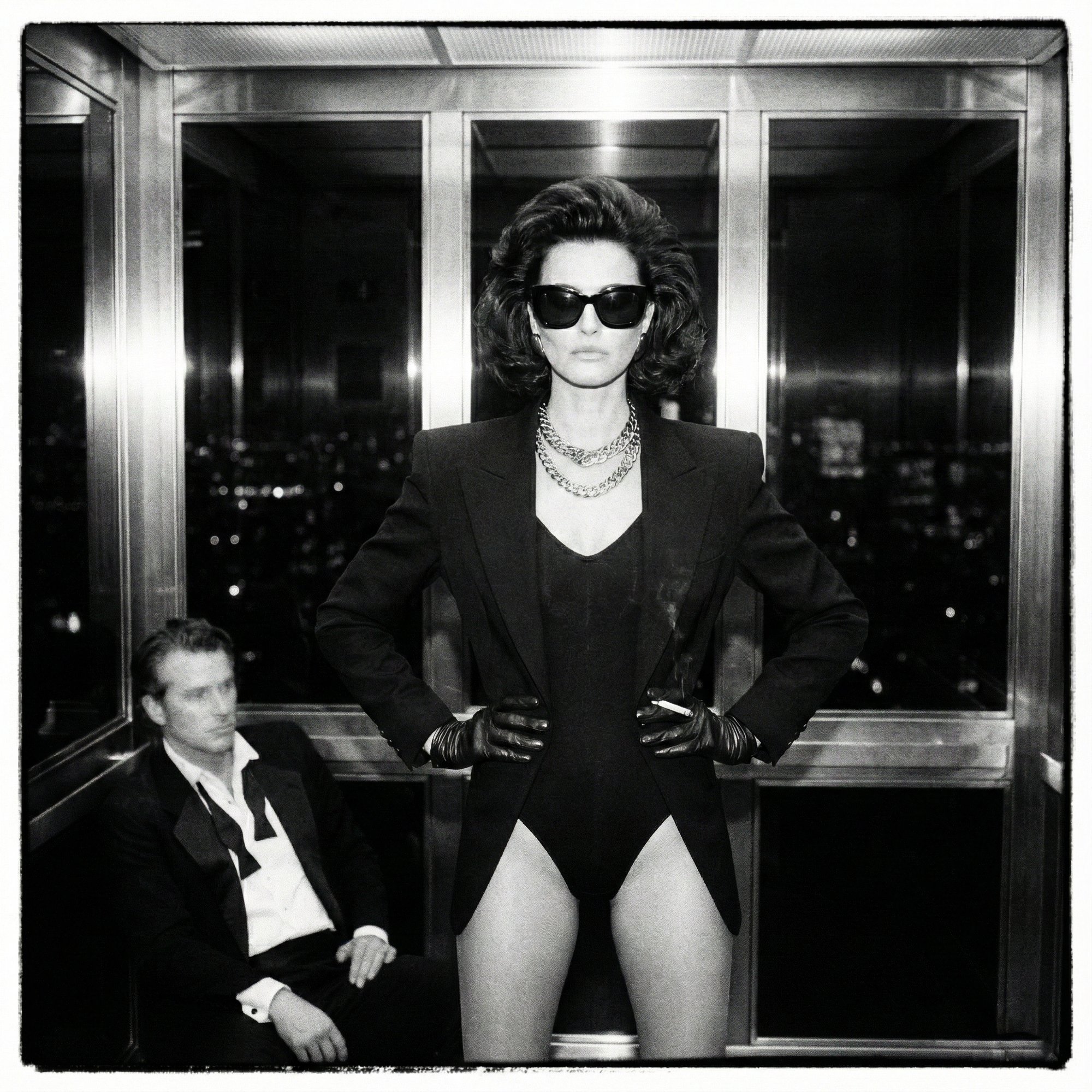 Black and white: a woman in a black bodysuit, blazer and sunglasses, hands on her hips in a mirrored elevator, a man in a tuxedo seated below