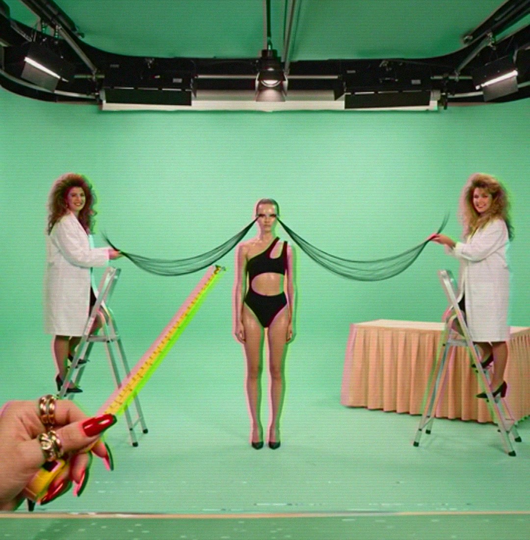 Ep. 02, Lengthening: the green studio under a lighting rig, two assistants in lab coats on ladders stretching a model’s lashes, a hand with red nails holding out a tape measure