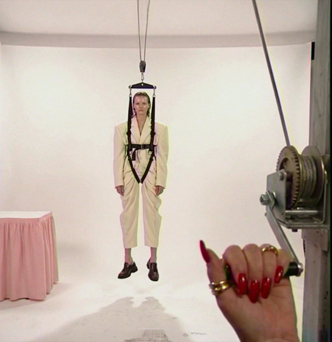 Ep. 01, Lifting: the white set, a model in a cream suit hanging in a harness from a cable, a pink skirted table, a hand with red nails and gold rings turning the winch