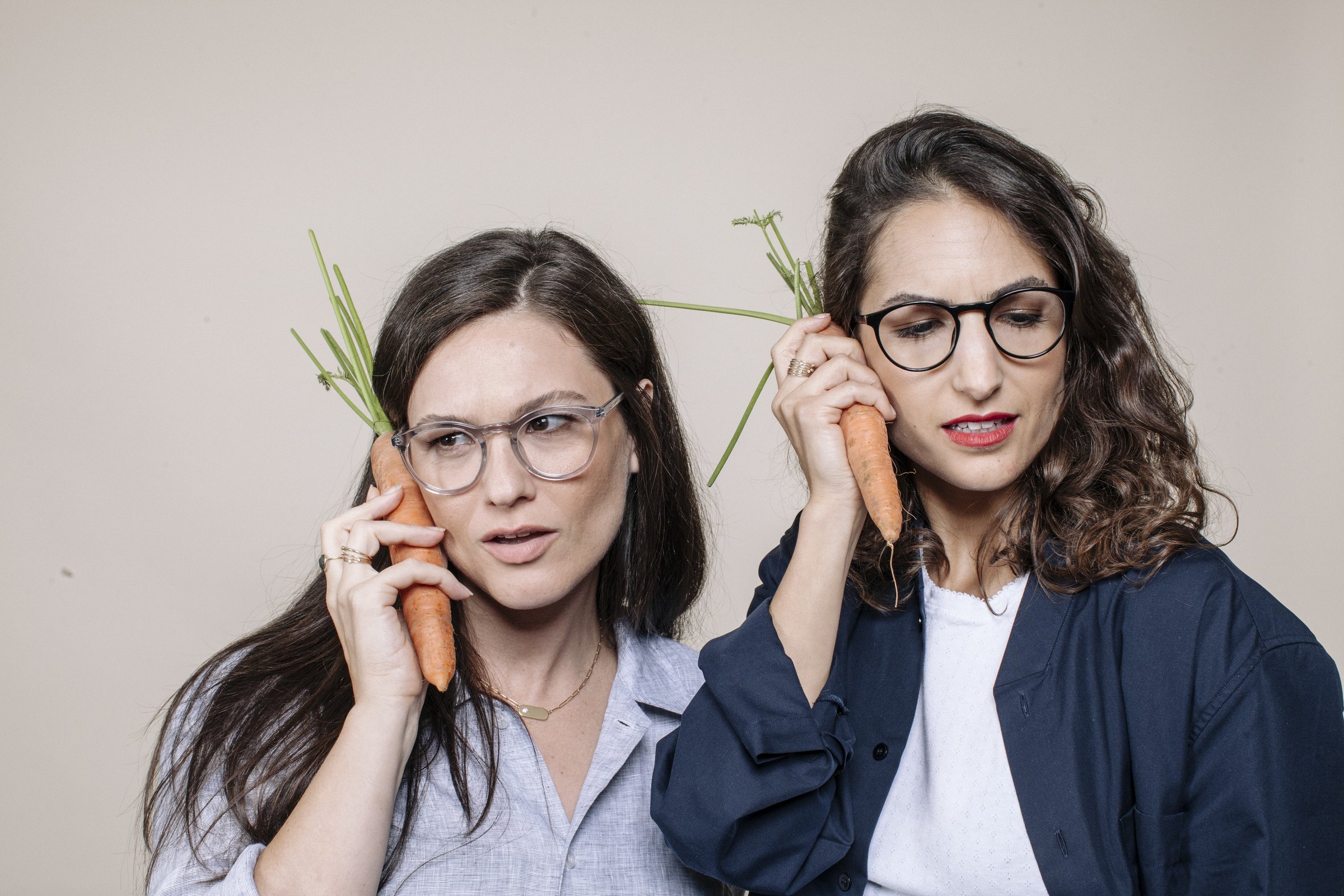 Two women in Zak. glasses holding carrots to their ears like phones, against a warm beige wall